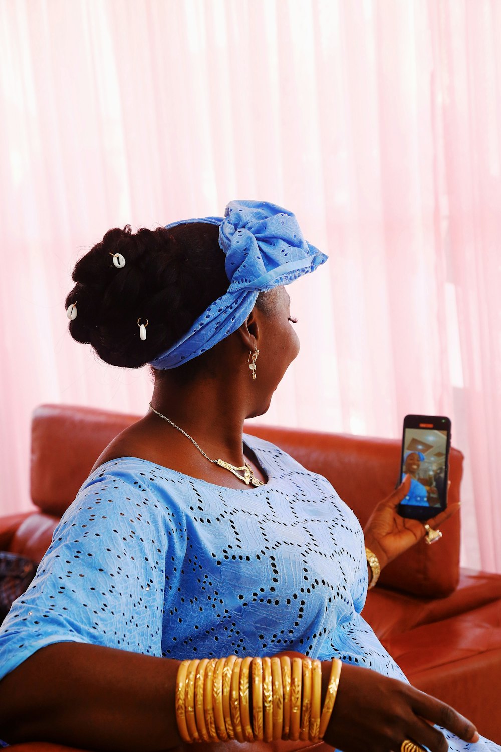 A smiling Nigerian woman in a blue lace dress and headwrap reading a message on her phone